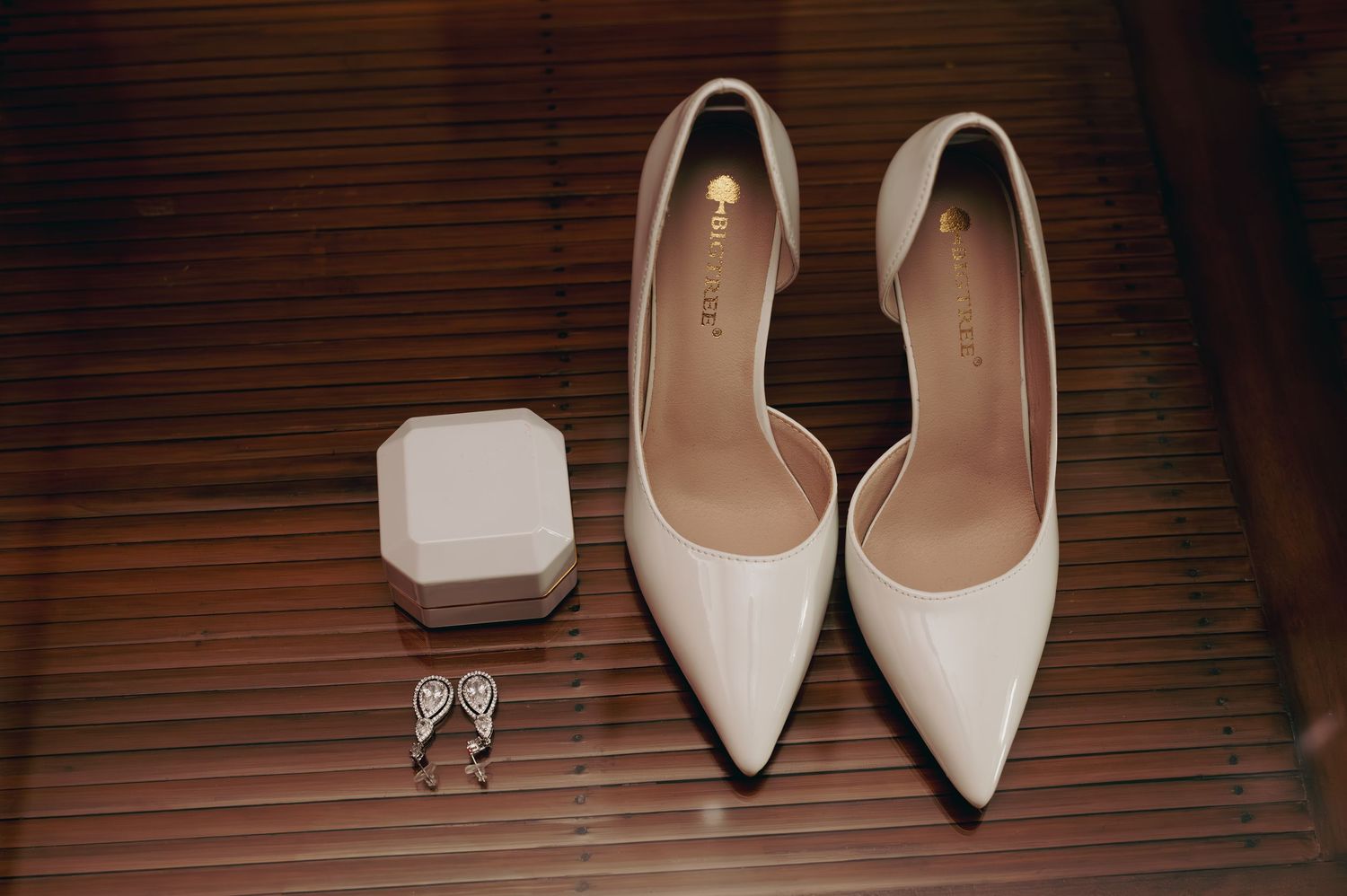 White pointed-toe bridal heels displayed with a ring box and delicate earrings on a bamboo surface.