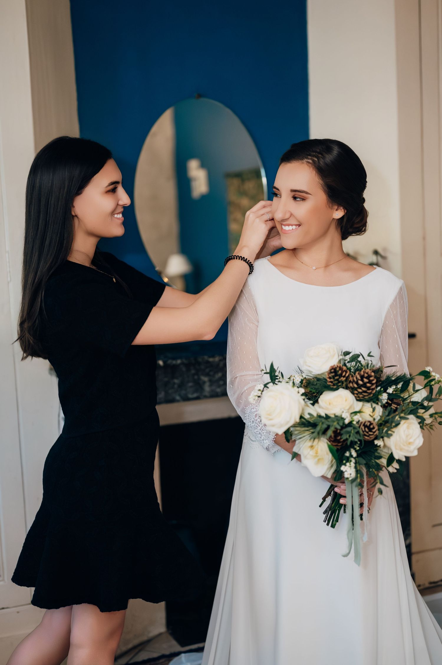 Woman in black dress gently touching bride's face while bride holds winter bouquet with white roses and pine cones.