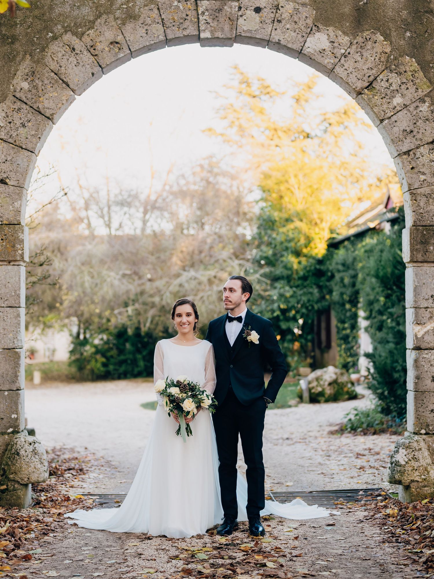 Bride and groom stand beneath a stone archway surrounded by autumn foliage at an outdoor wedding.