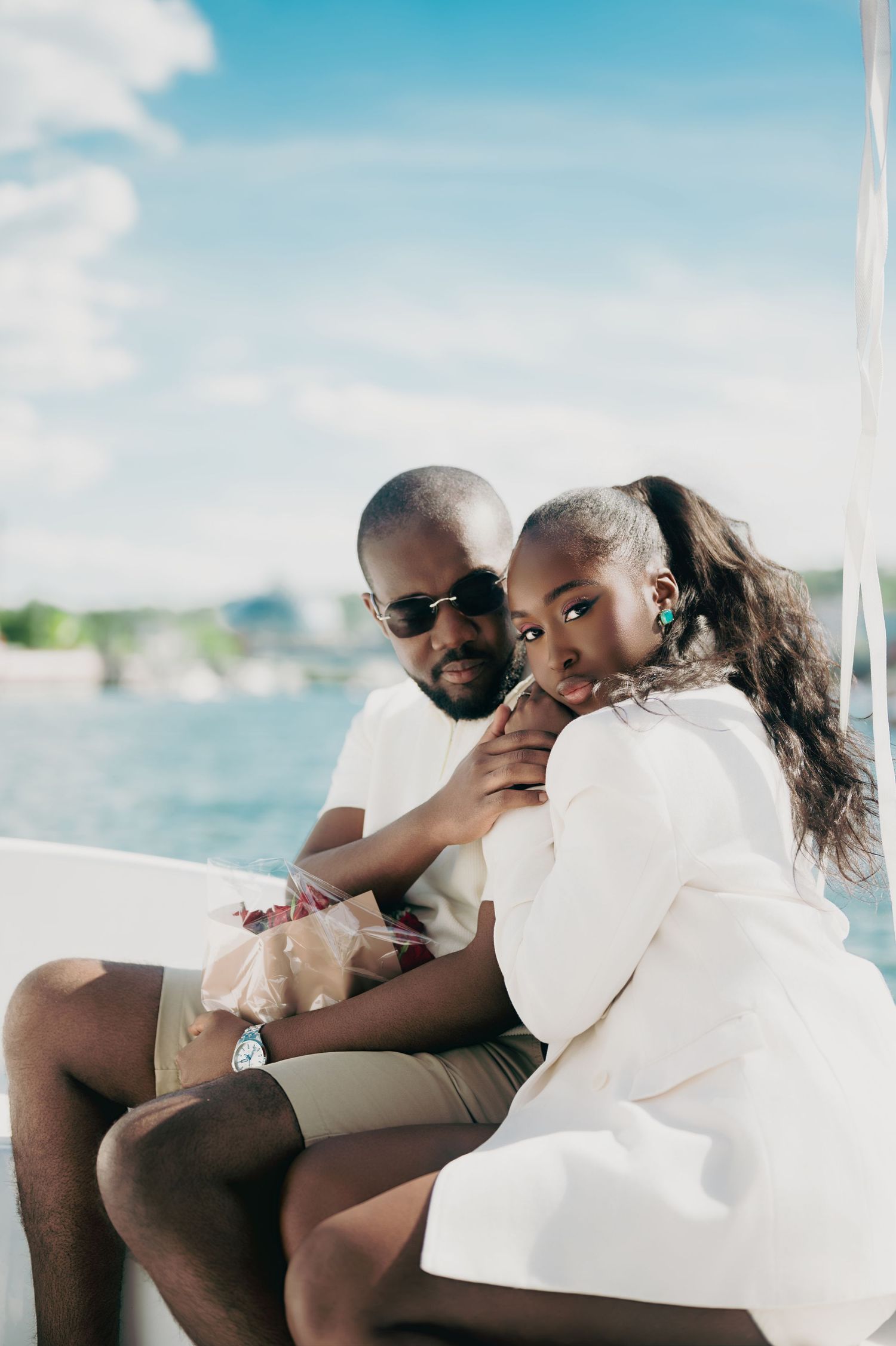 Stylish Black couple sitting together on a boat, woman in white blazer leaning against man in sunglasses.
