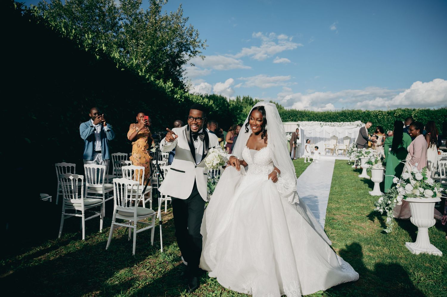 Bride in white ball gown walks with groom at outdoor wedding reception on green lawn.