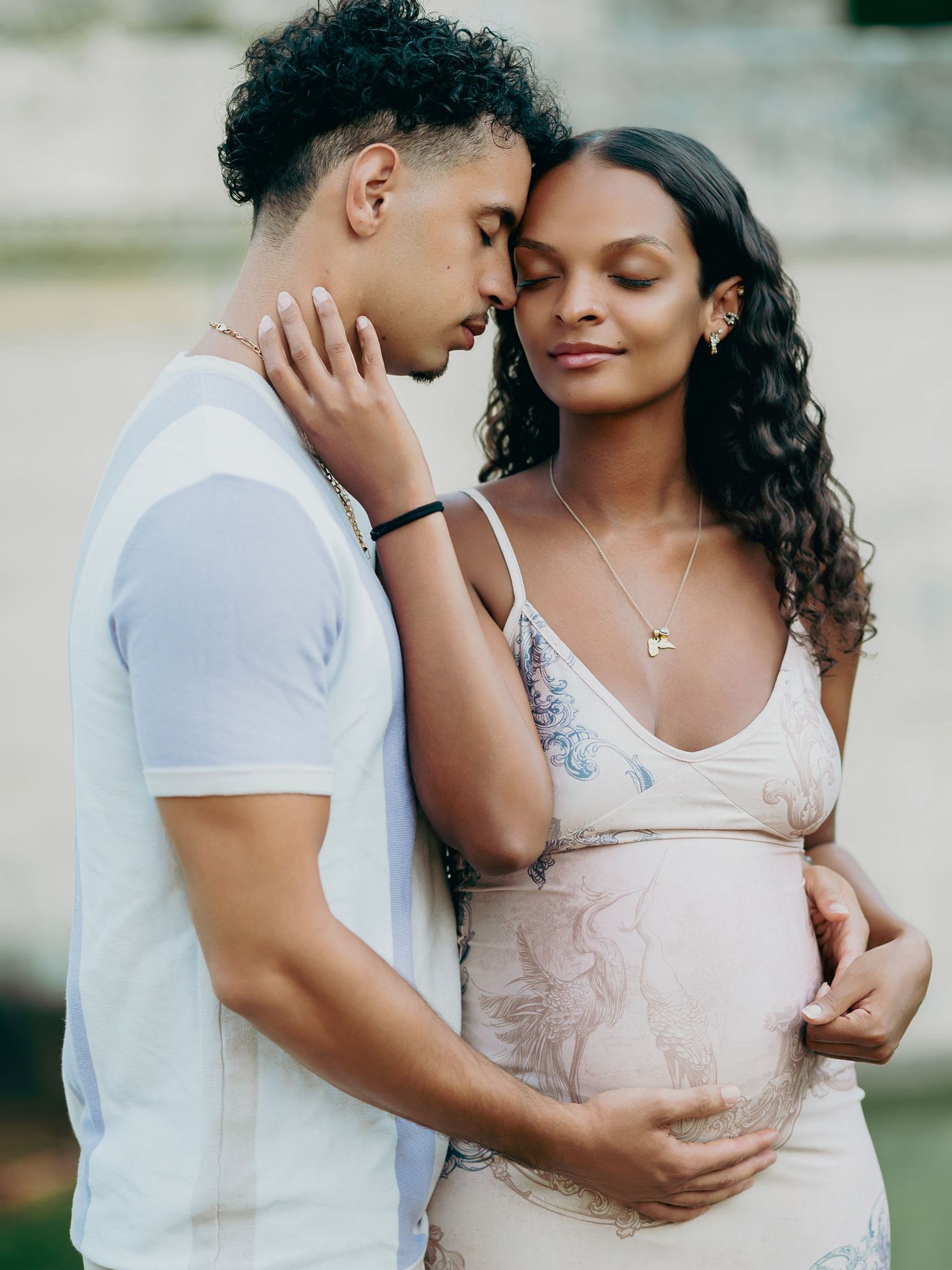 Couple sharing an intimate forehead-to-forehead moment while gently cradling a baby bump outdoors.