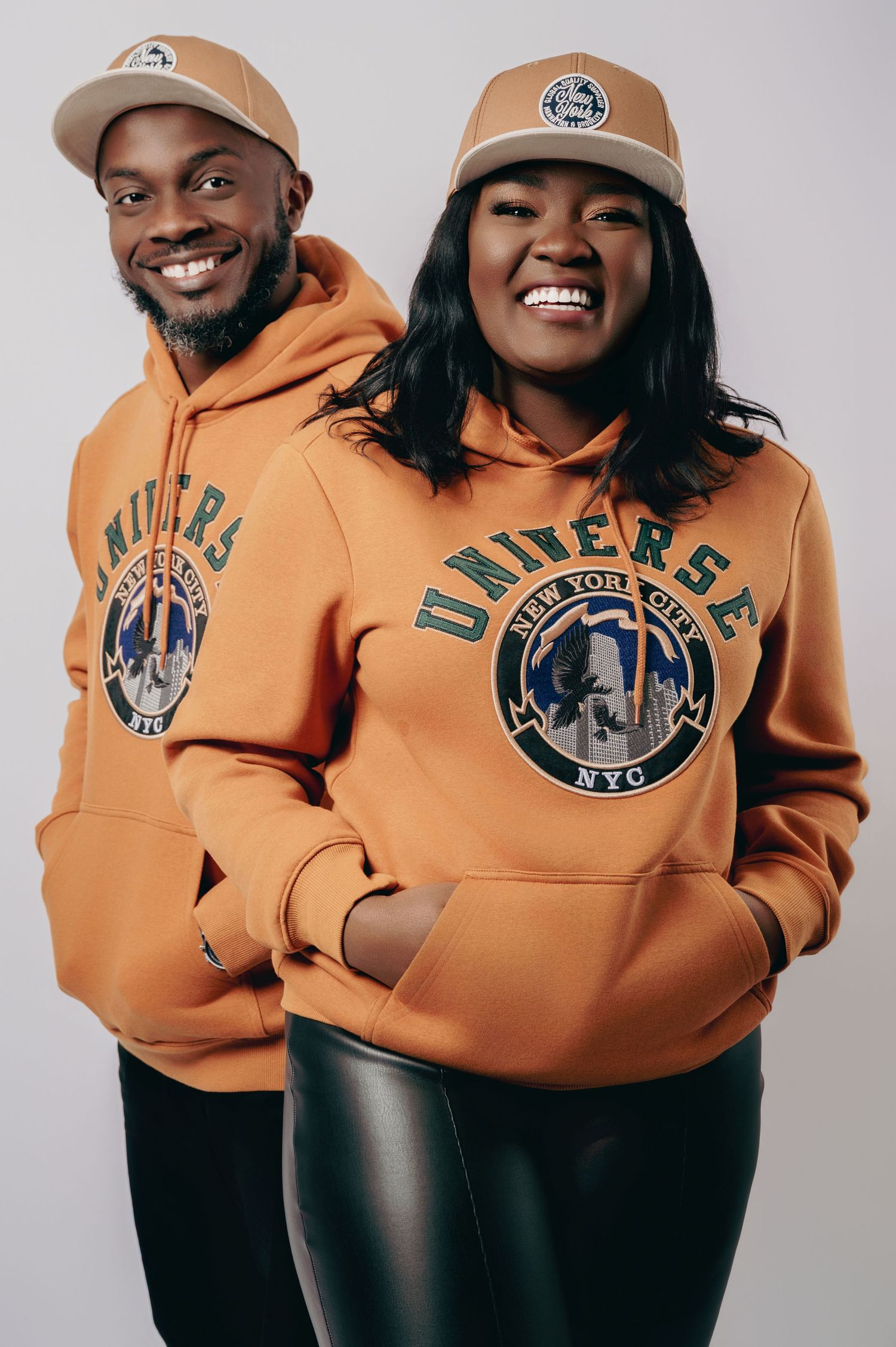 Smiling Black couple wearing matching orange Universe NYC hoodies and tan snapback caps.