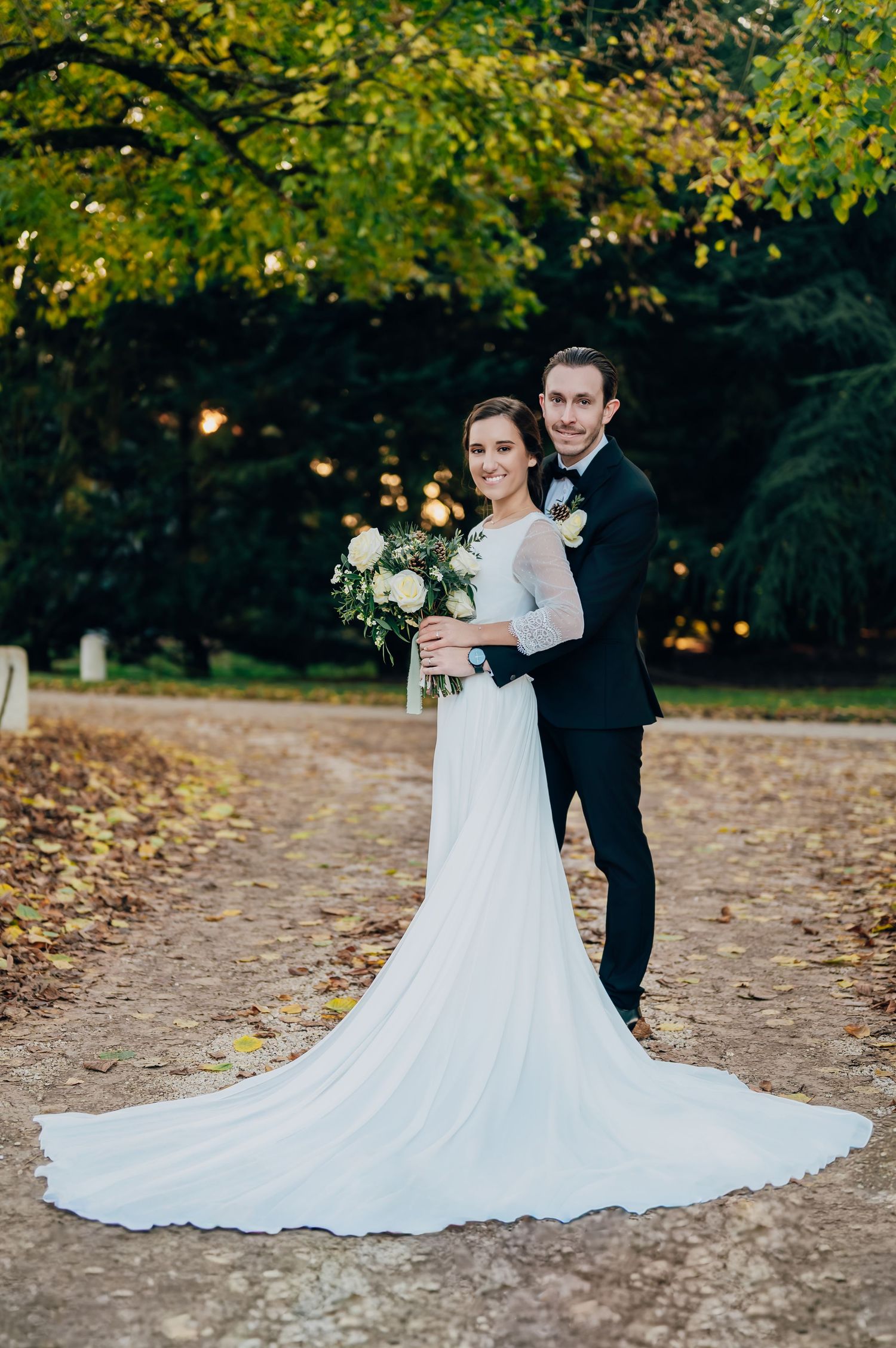 Happy bride and groom posing on a tree-lined autumn path, bride in a long-train white gown.