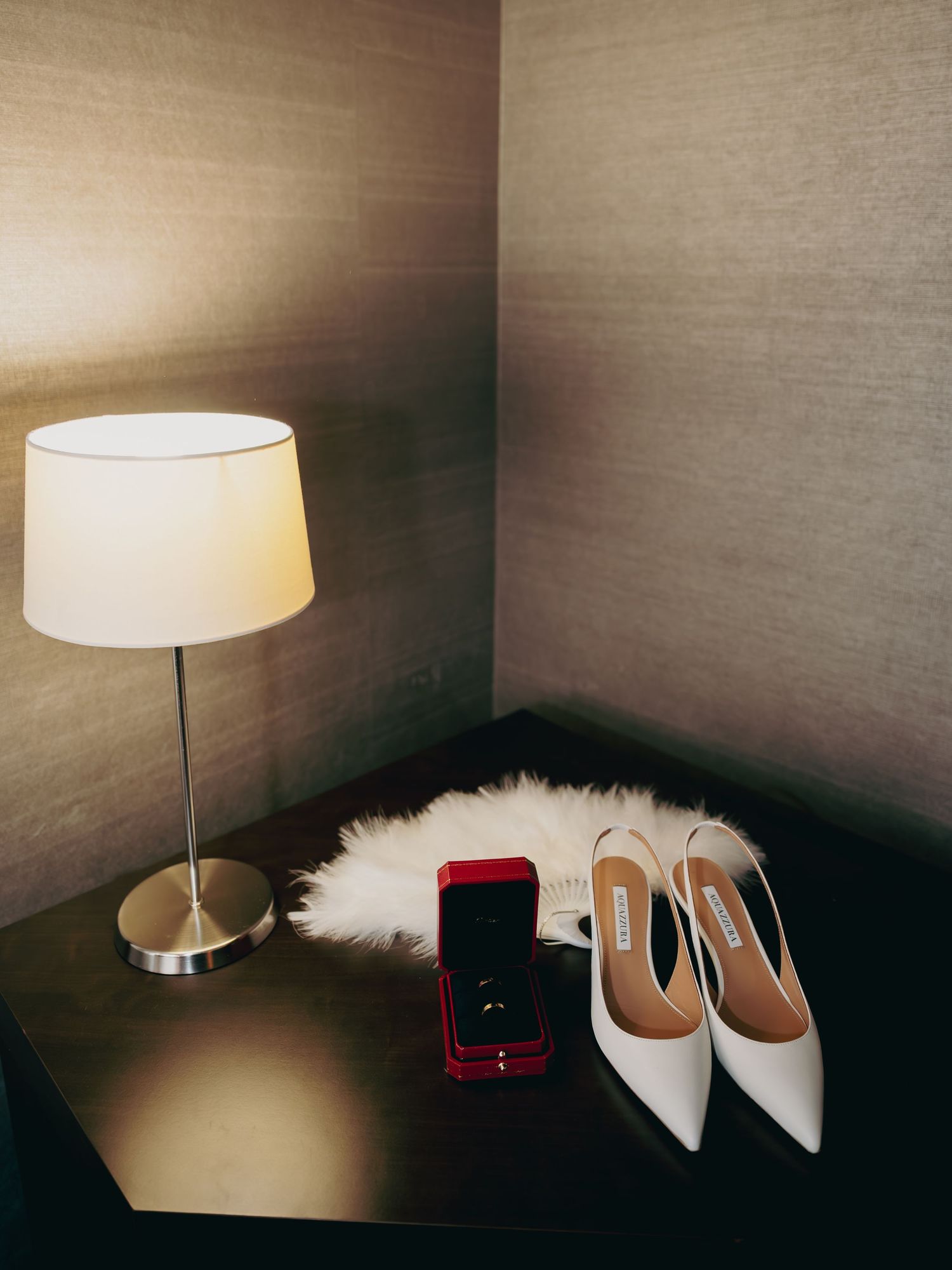 White bridal heels and red ring box displayed on a fur mat beside a glowing table lamp.