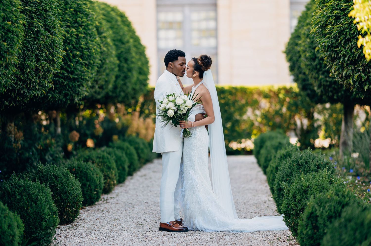 Bride and groom share a romantic moment on a gravel garden path lined with trimmed hedges and topiaries.