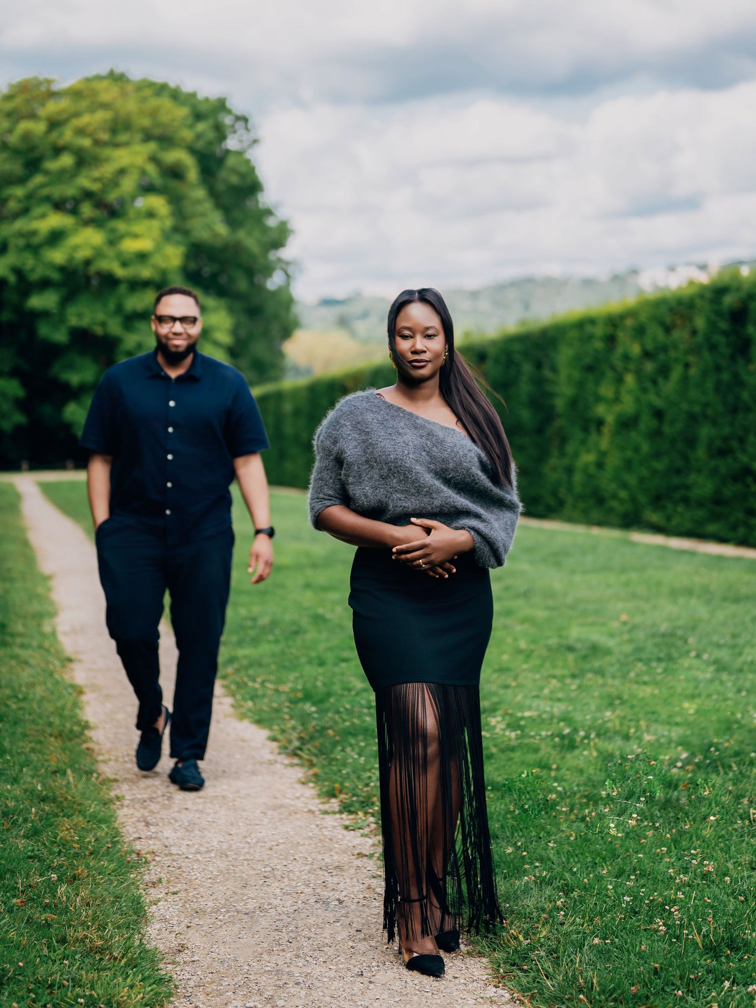 Pregnant woman in gray wrap and black fringe skirt walks confidently on garden path with man behind her.