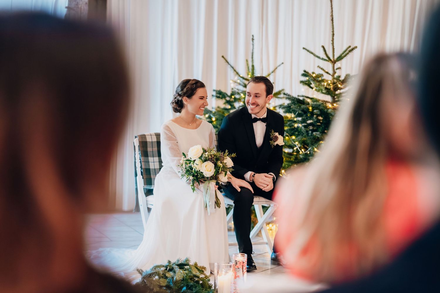 Bride and groom smiling during winter wedding ceremony with Christmas trees in decorated barn venue.