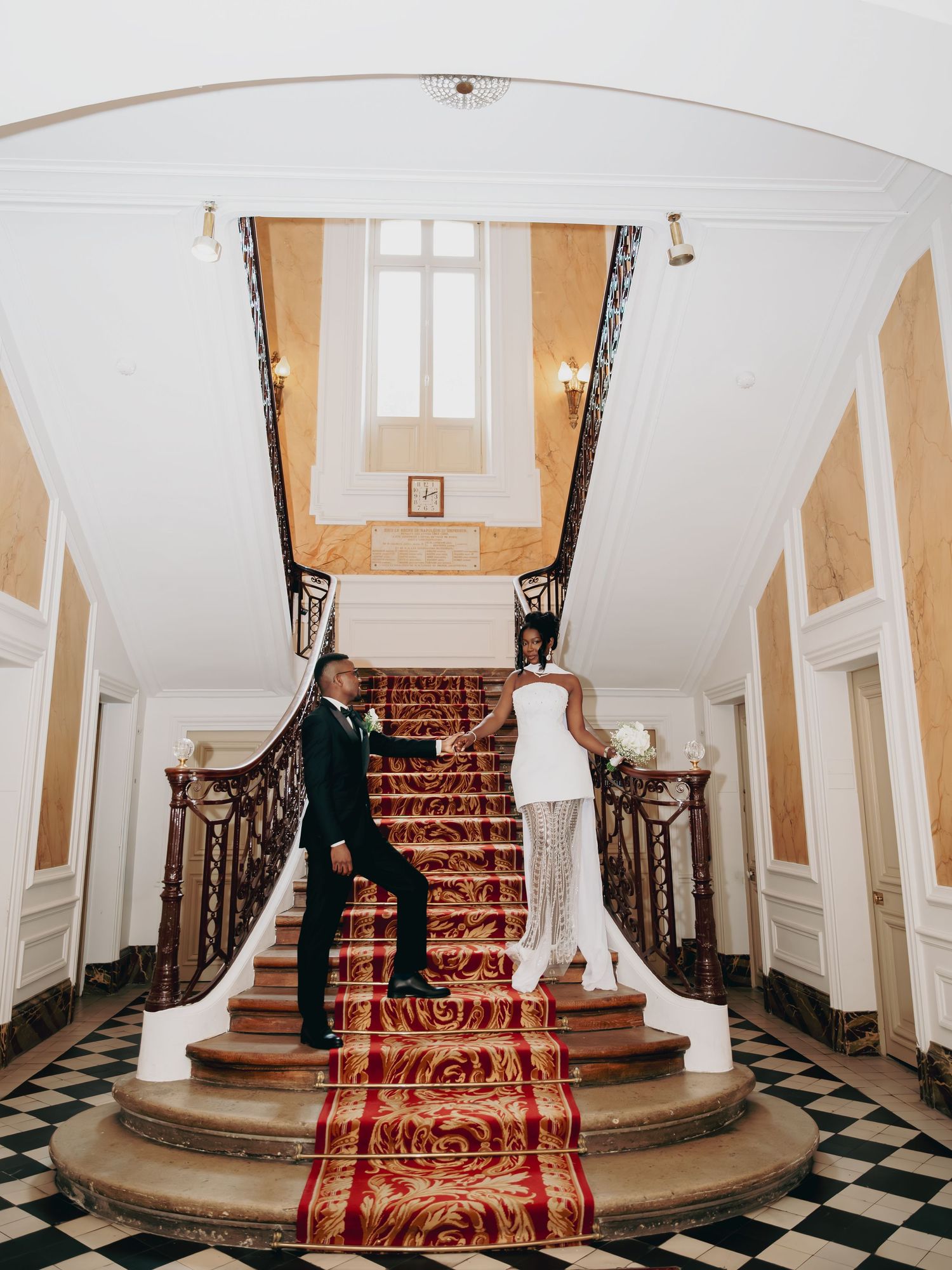 Bride and groom holding hands descending a grand staircase with red carpet in elegant venue.