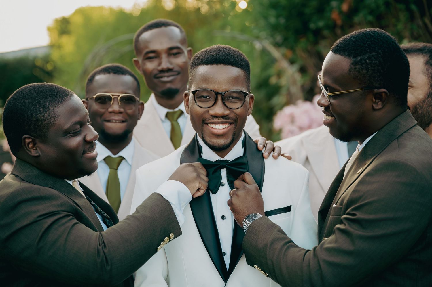 Smiling groom in white tuxedo surrounded by groomsmen adjusting his bow tie and suit.