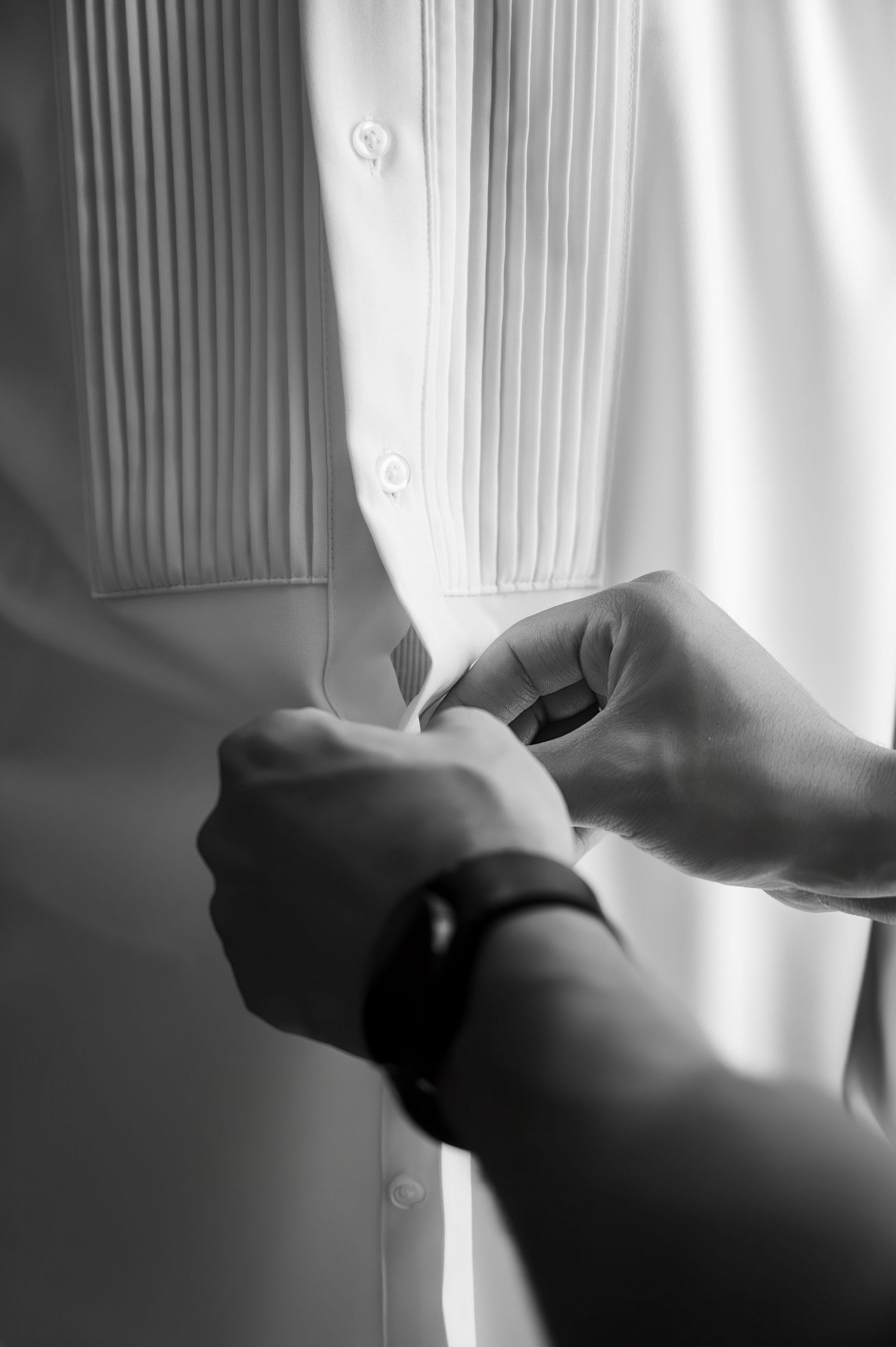 Black and white close-up of hands fastening cufflinks on grooms pleated dress shirt.