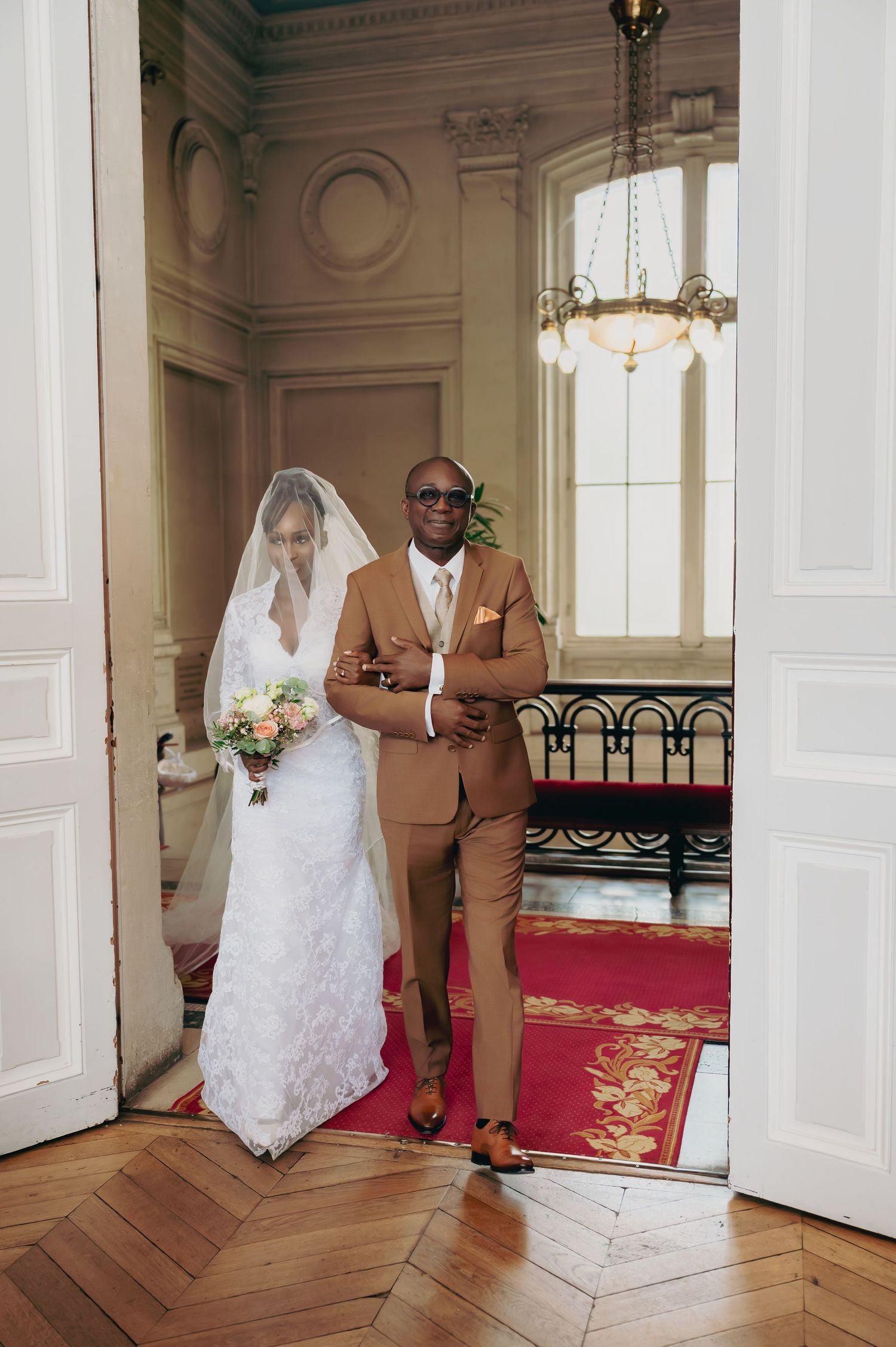 Bride in lace gown and veil poses with escort in tan suit at venue entrance.