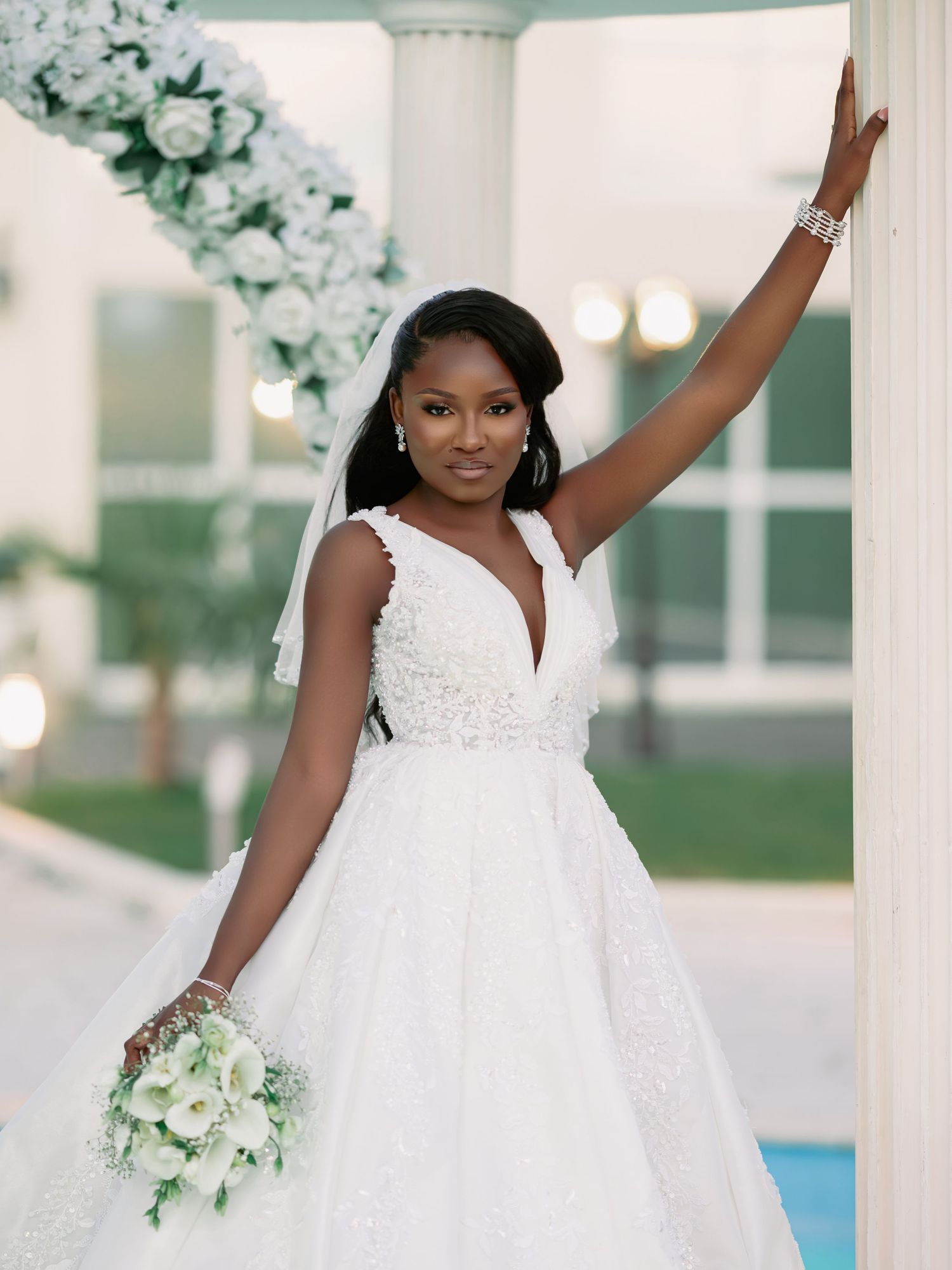 Beautiful Black bride in white ball gown posing by a column with white floral arrangements.