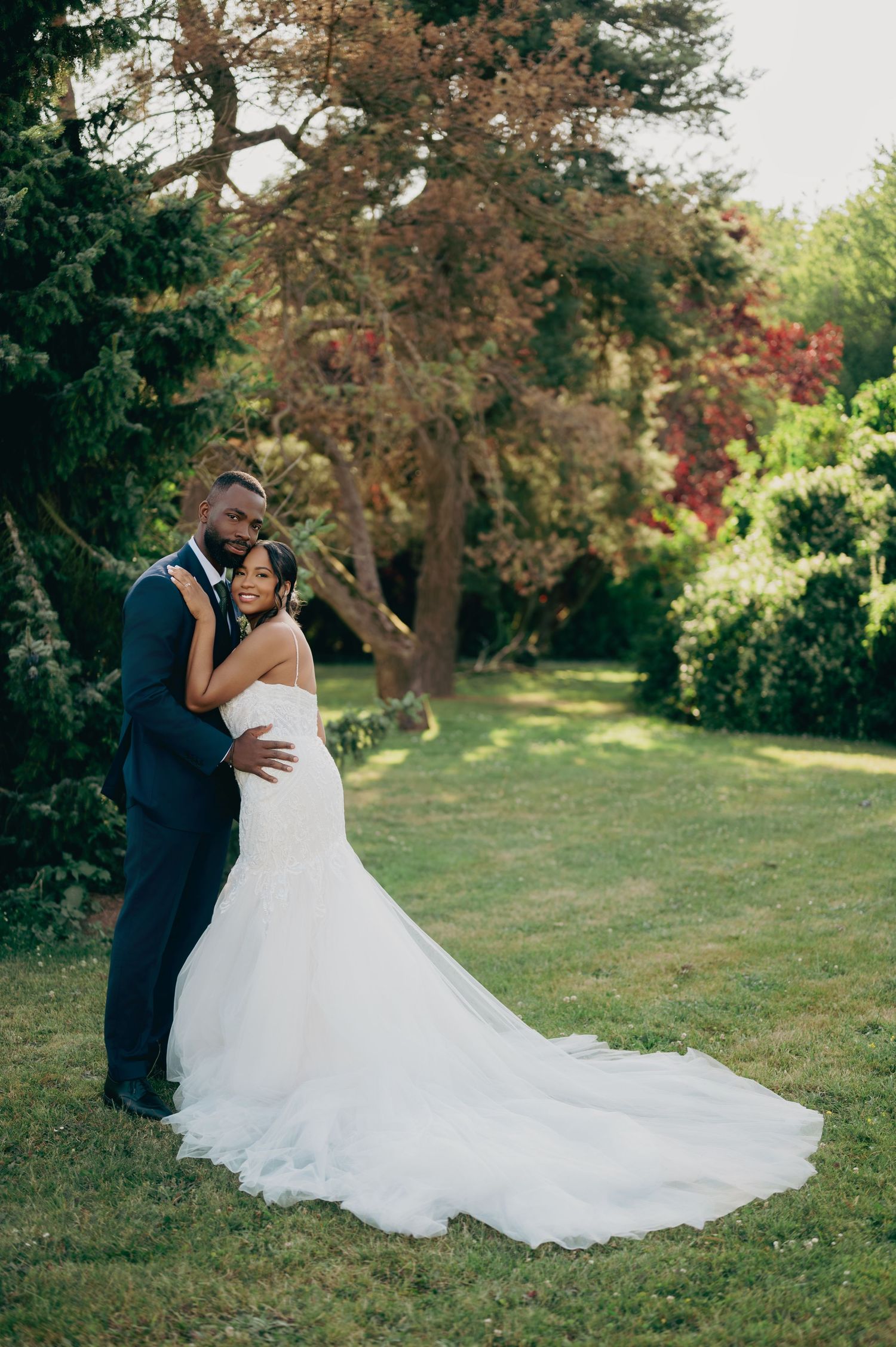 Groom embraces bride in fitted white gown with long train on lush green garden lawn at wedding.