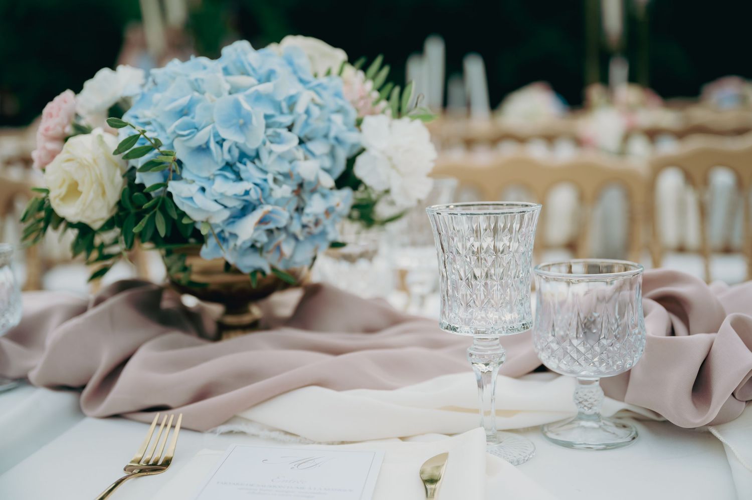 Wedding table centerpiece with blue hydrangeas, crystal goblets, and dusty rose linen runner.