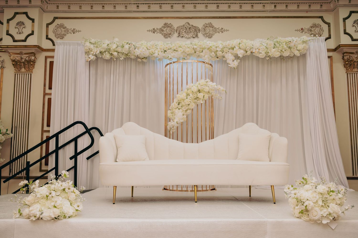 Elegant wedding stage with white sofa, draped backdrop, and white floral arch decor.