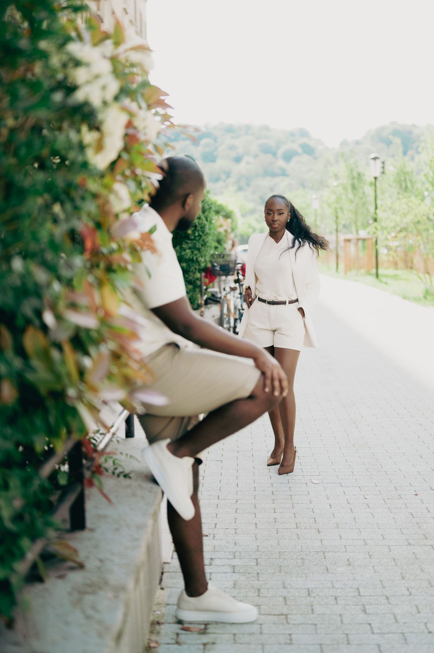 A couple in white outfits interact casually on a cobblestone path beside a lush green hedge.