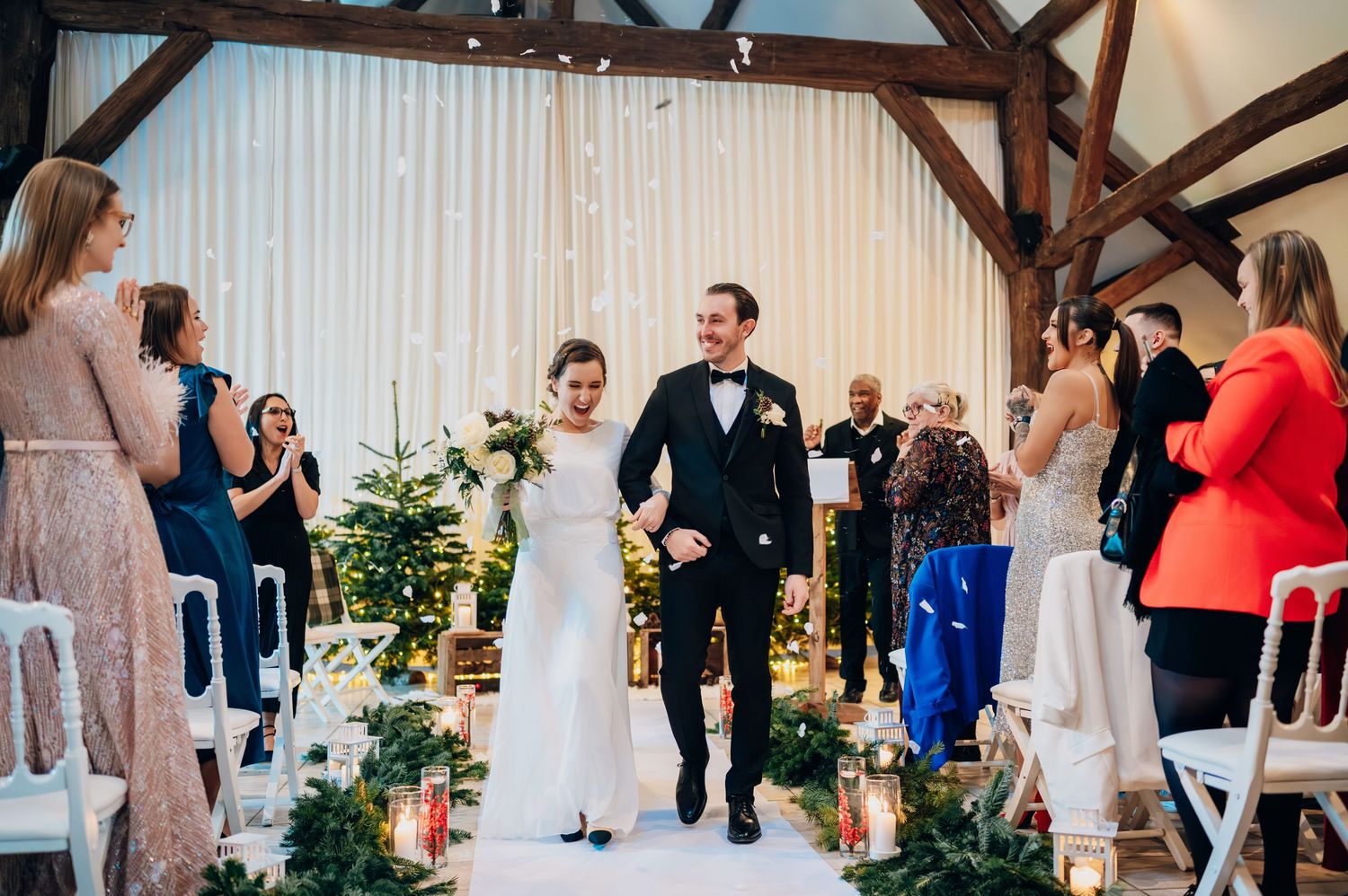 Happy bride and groom walk down decorated aisle lined with greenery and candles.