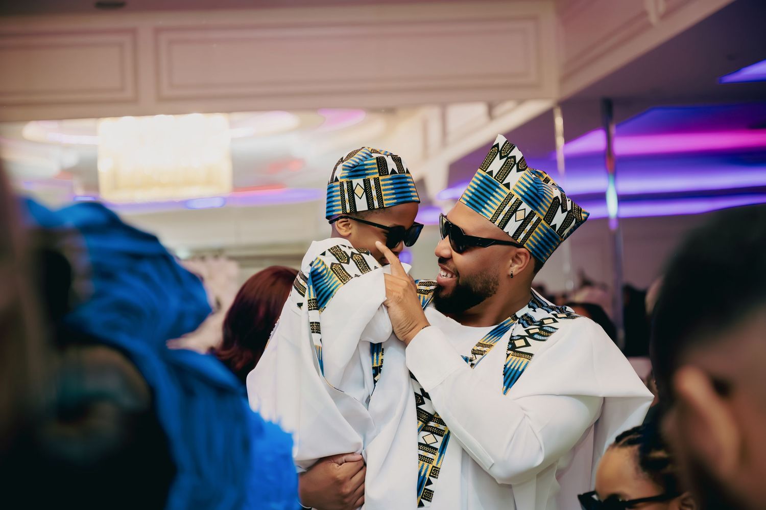 Man in white African agbada outfit holds a baby dressed in matching traditional attire.