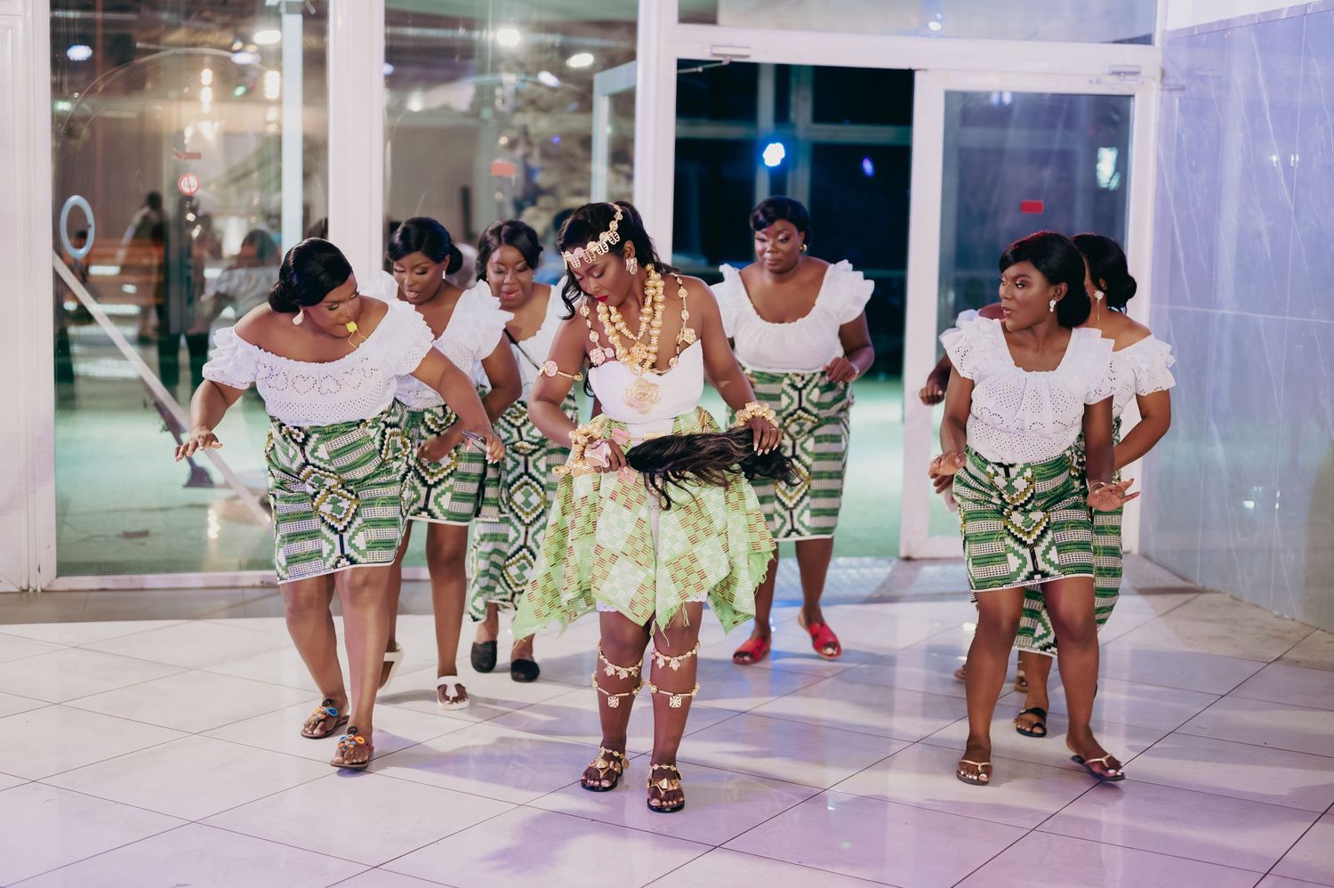 Women in matching white tops and green printed skirts perform a traditional African dance at a wedding celebration.