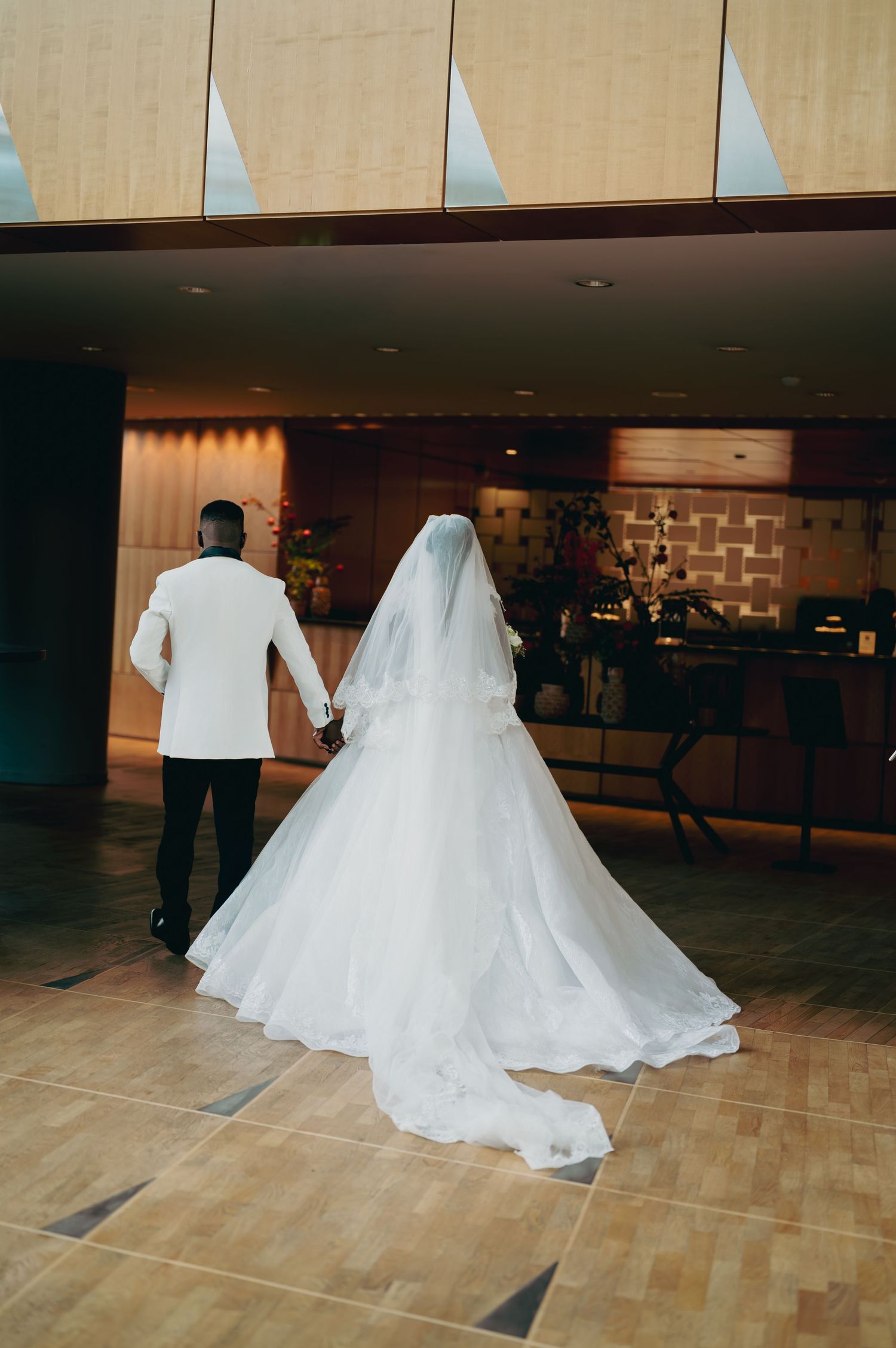 Black bride and groom walking hand in hand, bride's long white veil trailing behind in hotel lobby.