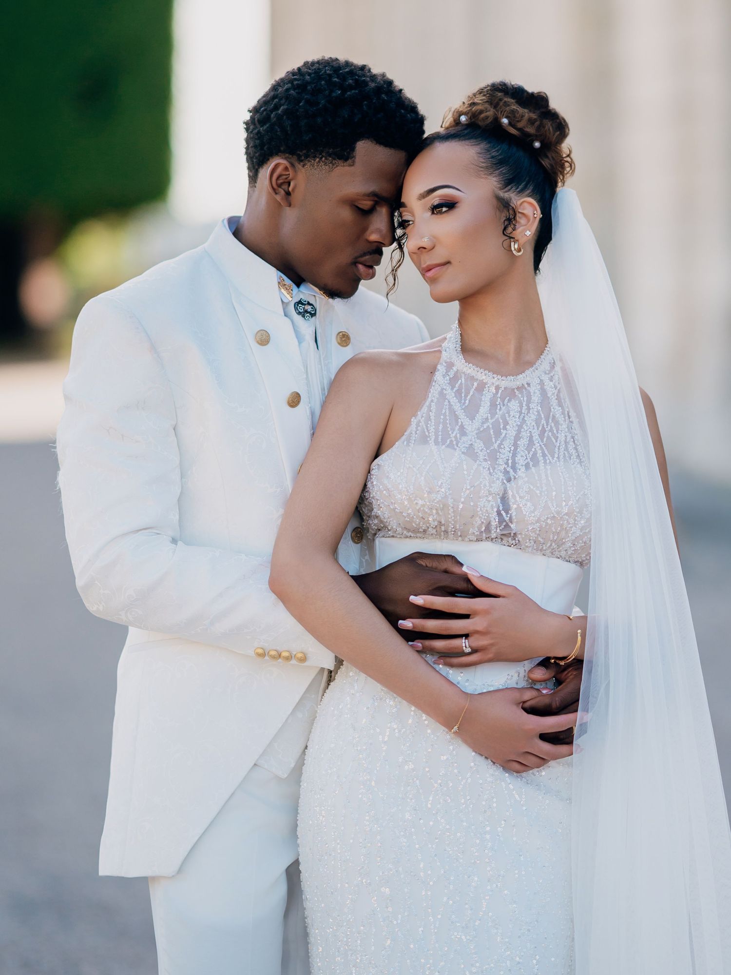 A couple in wedding attire embrace tenderly, groom in white suit, bride in lace gown with veil.