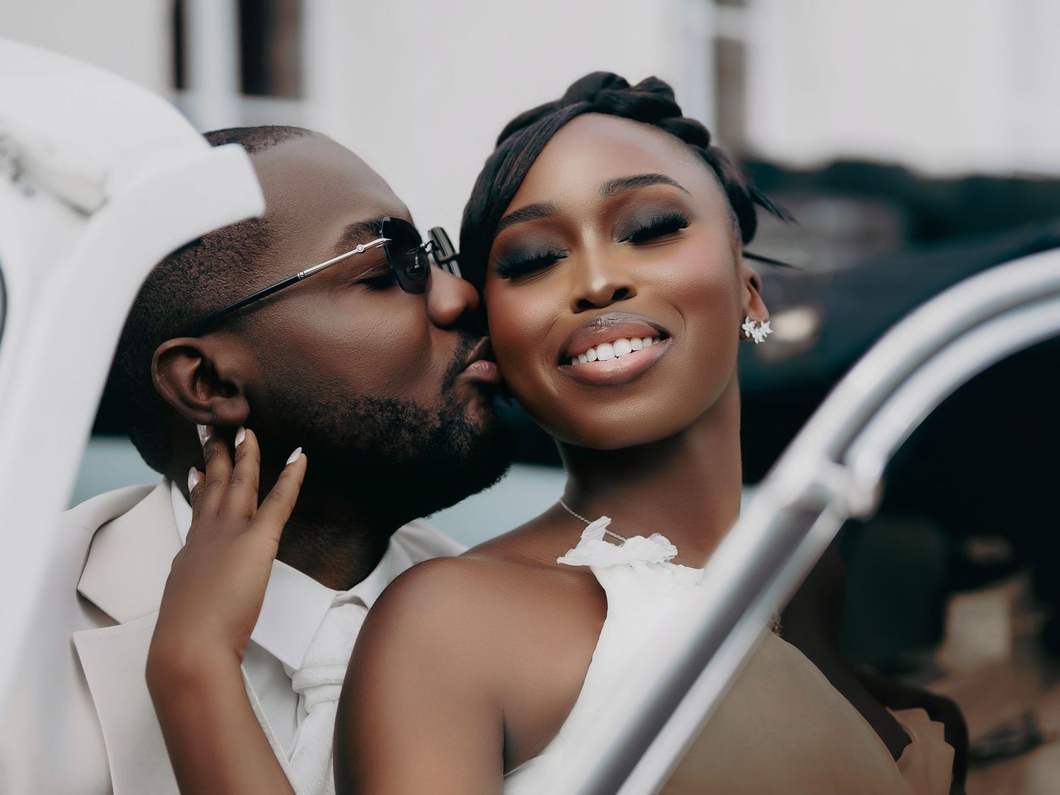 Groom in white suit kisses bride on cheek as she smiles joyfully near a white vintage car.