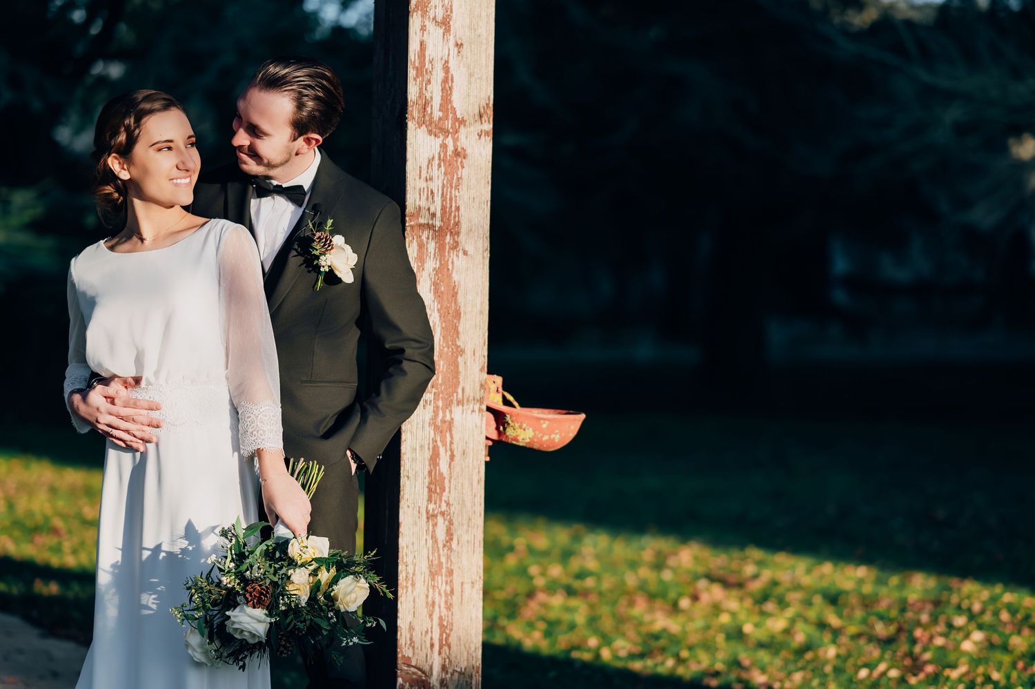Bride and groom pose beside a wooden post in a sunlit park, bride holding lush floral bouquet.