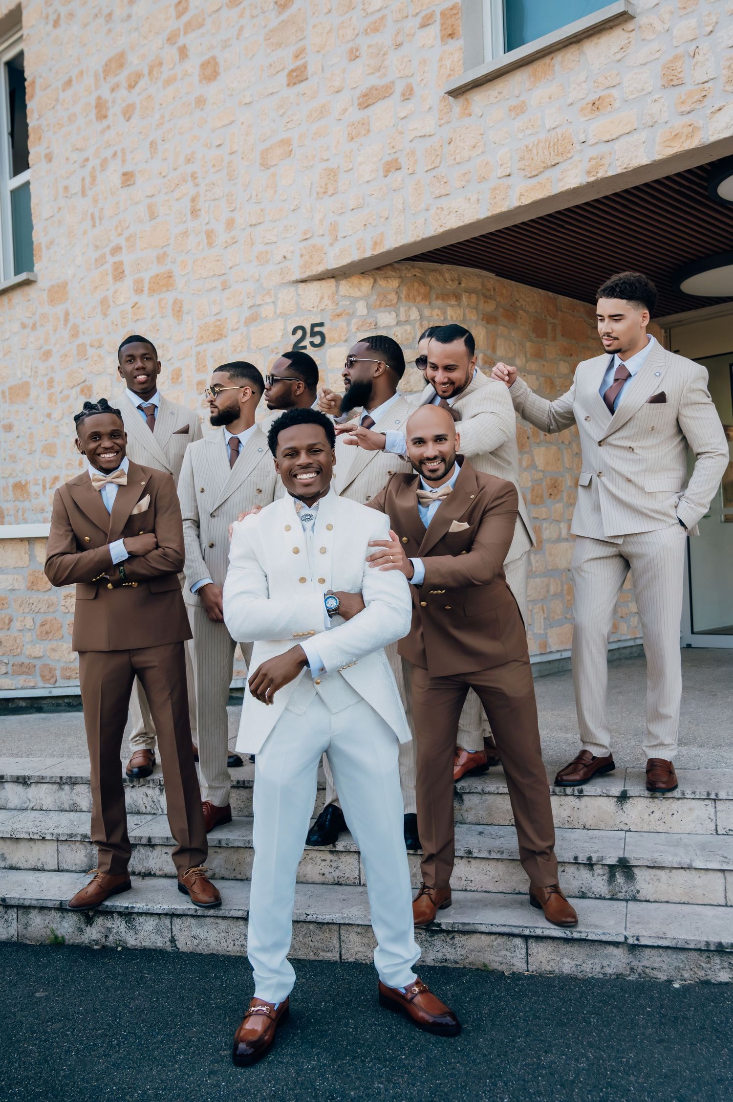 Smiling groomsmen in brown and beige suits laughing on steps with groom in white suit.