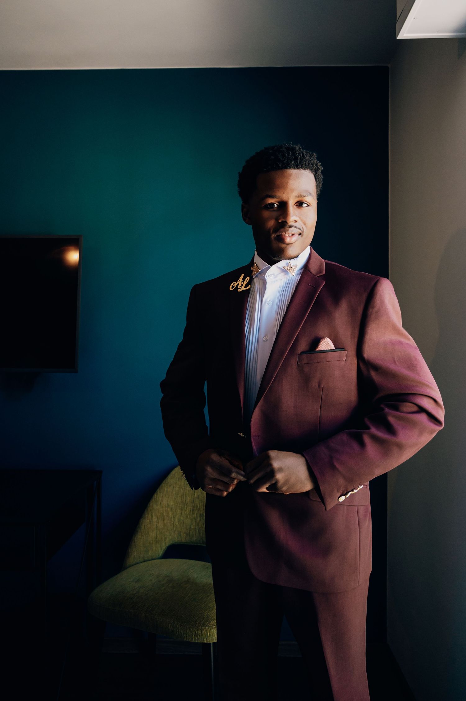 Dapper groom posing in a burgundy double-breasted suit with white shirt and pocket square in a stylish hotel room.