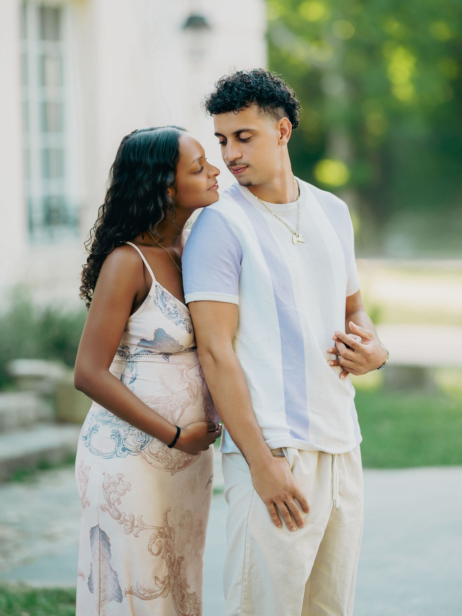 Pregnant couple in soft floral dress and white tee embracing outdoors in warm natural light.