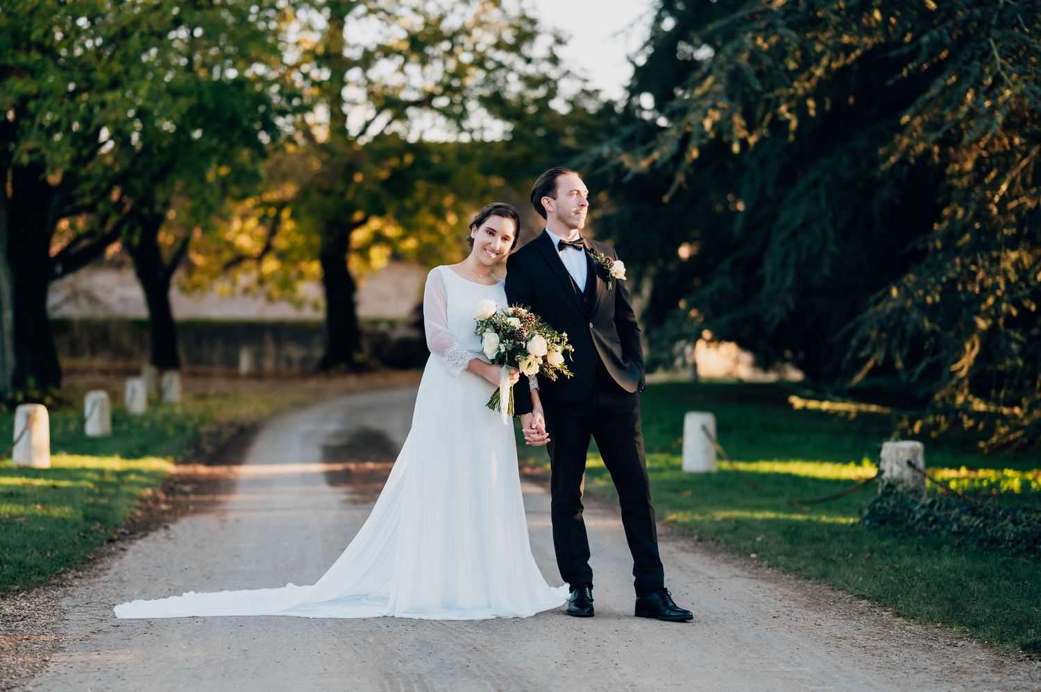 Elegant bride and groom pose on tree-lined path during golden hour wedding portraits.