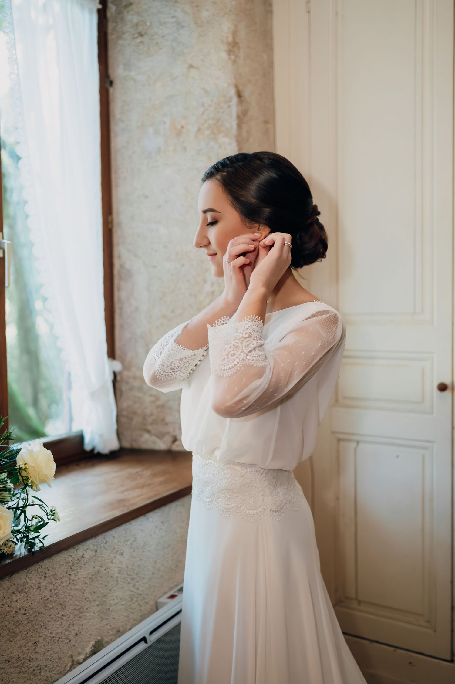 Elegant bride in lace wedding gown putting on earrings by a window before ceremony.