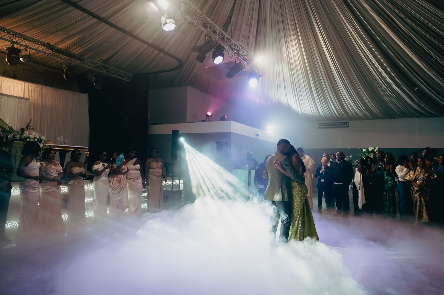 Couple shares first dance surrounded by dramatic fog effects under spotlights at wedding reception.