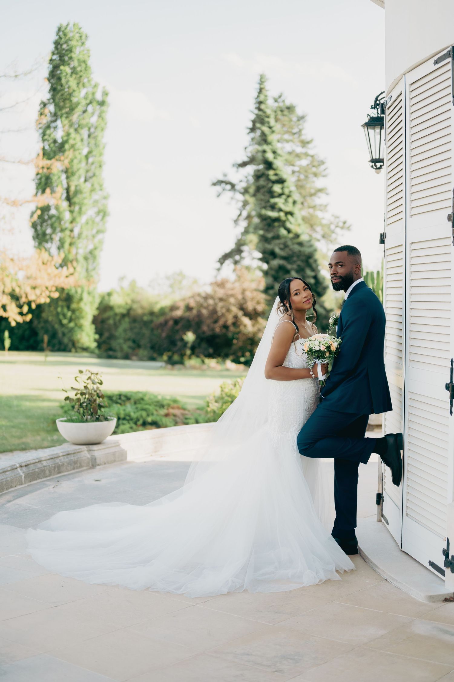 Bride in a flowing white gown with long veil stands with groom in navy suit at a chateau.