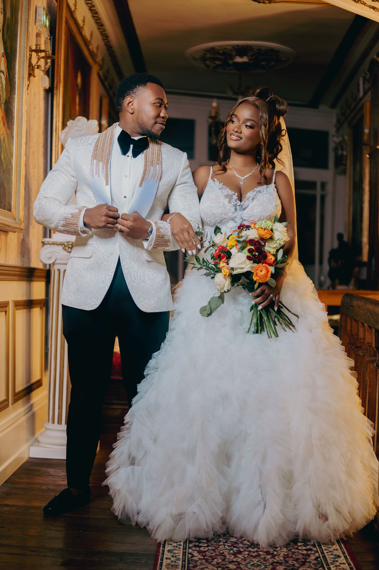 Bride and groom posing in ornate venue, groom in white tuxedo, bride in ruffled ball gown with bouquet.