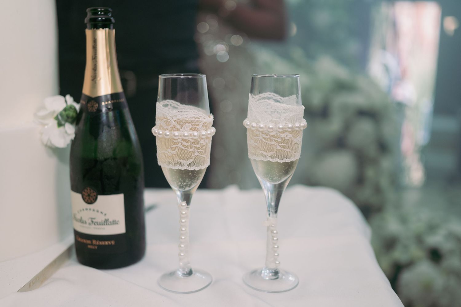 Two champagne flutes decorated with lace and pearls beside a bottle of champagne at a wedding reception.
