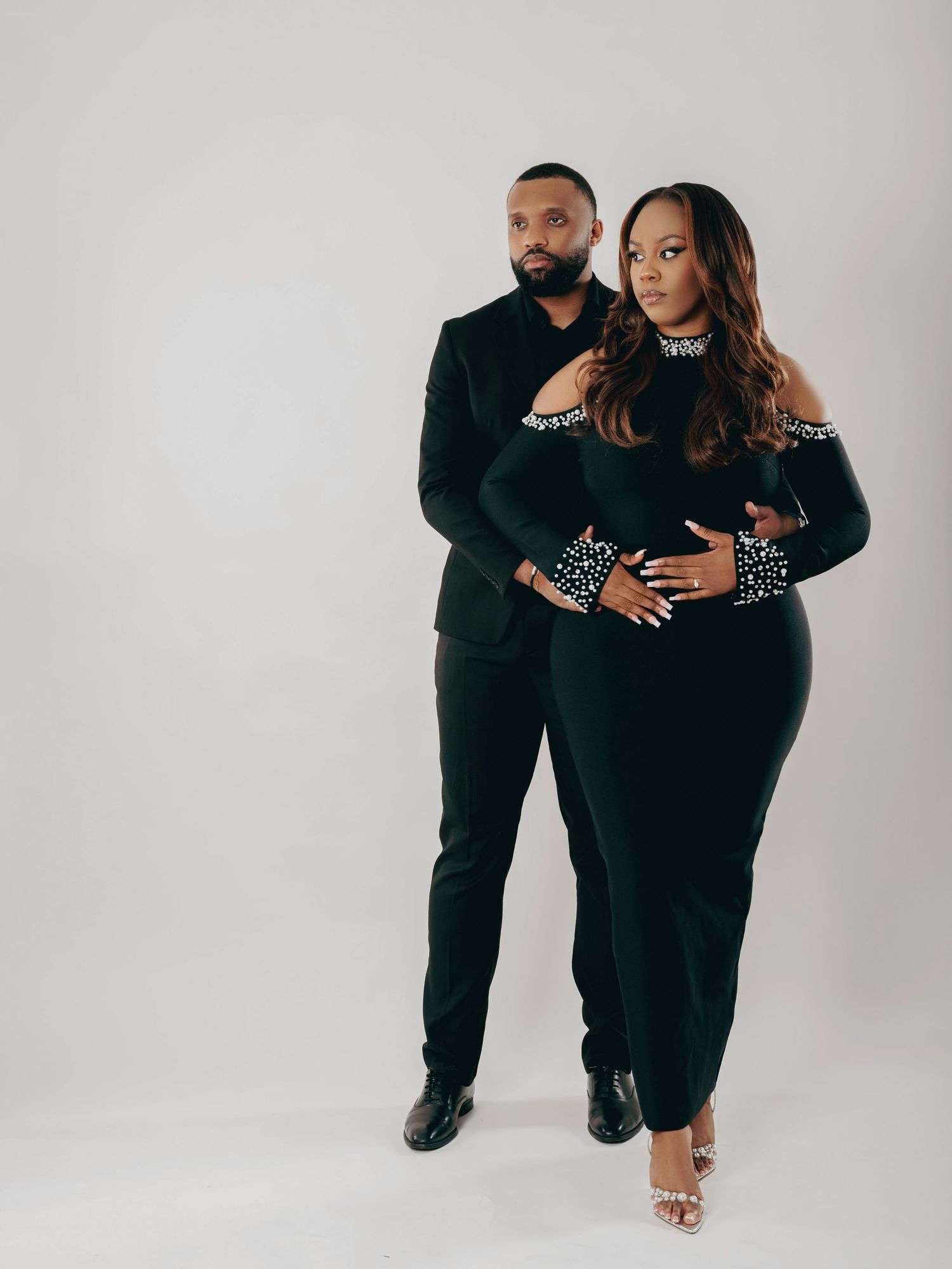 Elegant couple in matching black pearl-embellished outfits posing for a maternity photo shoot.