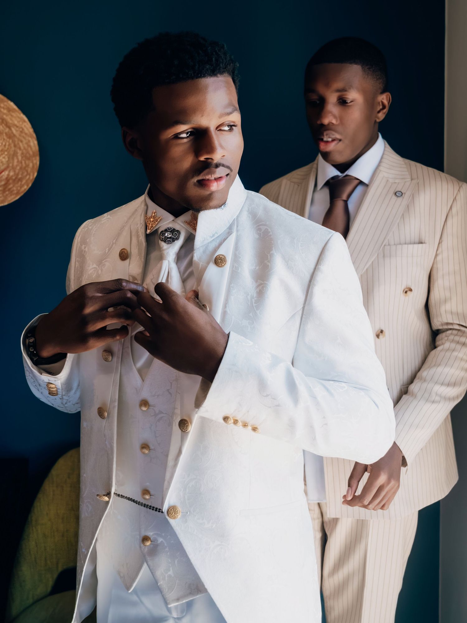Stylish groom in white double-breasted suit with gold buttons and bow tie prepares with groomsman behind him.