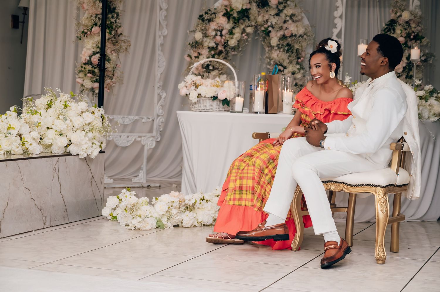 Couple in traditional Caribbean wedding attire sitting together at elegant floral decorated reception venue.
