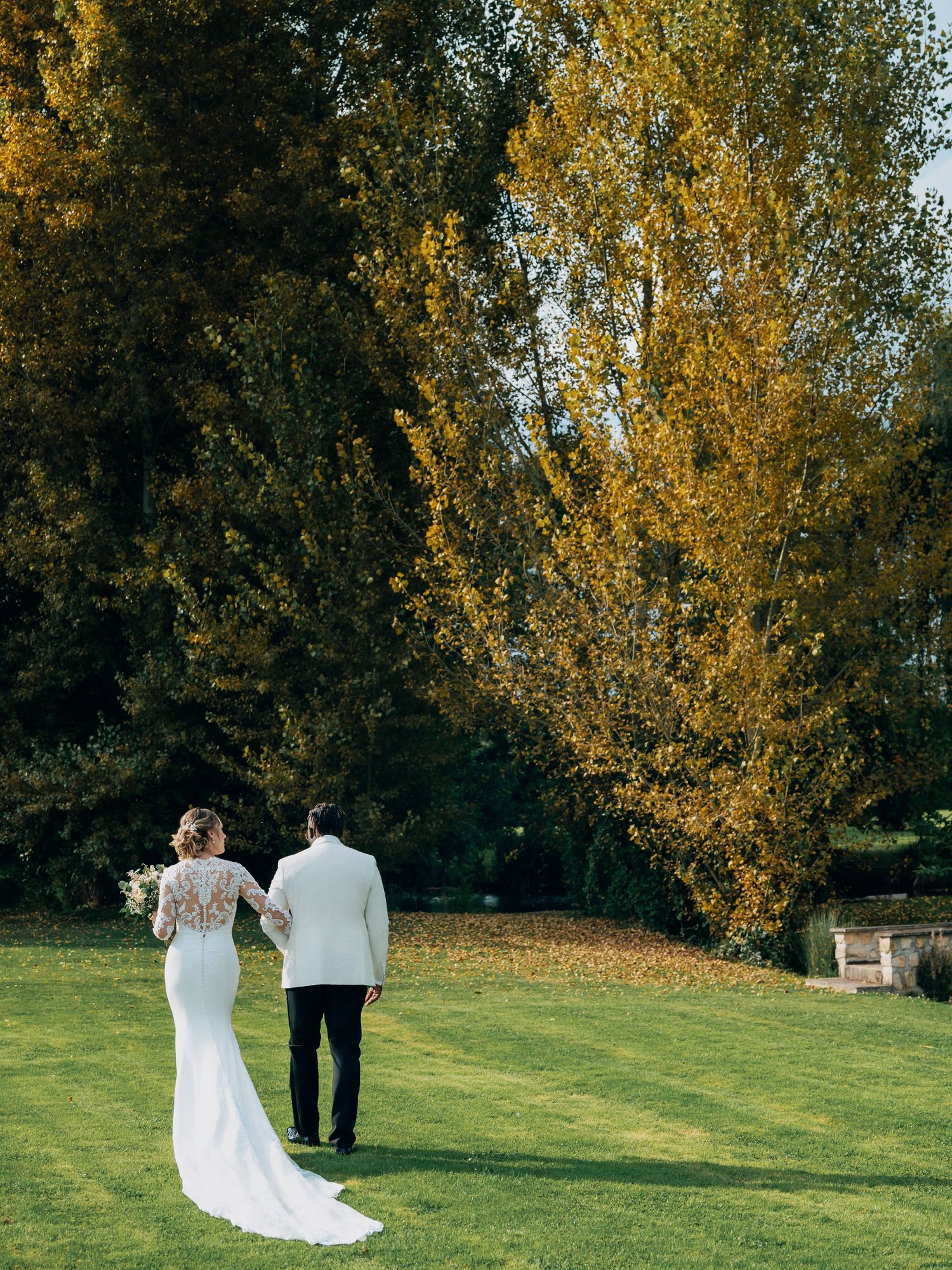 Bride and groom walking hand in hand across a green lawn surrounded by golden autumn trees.