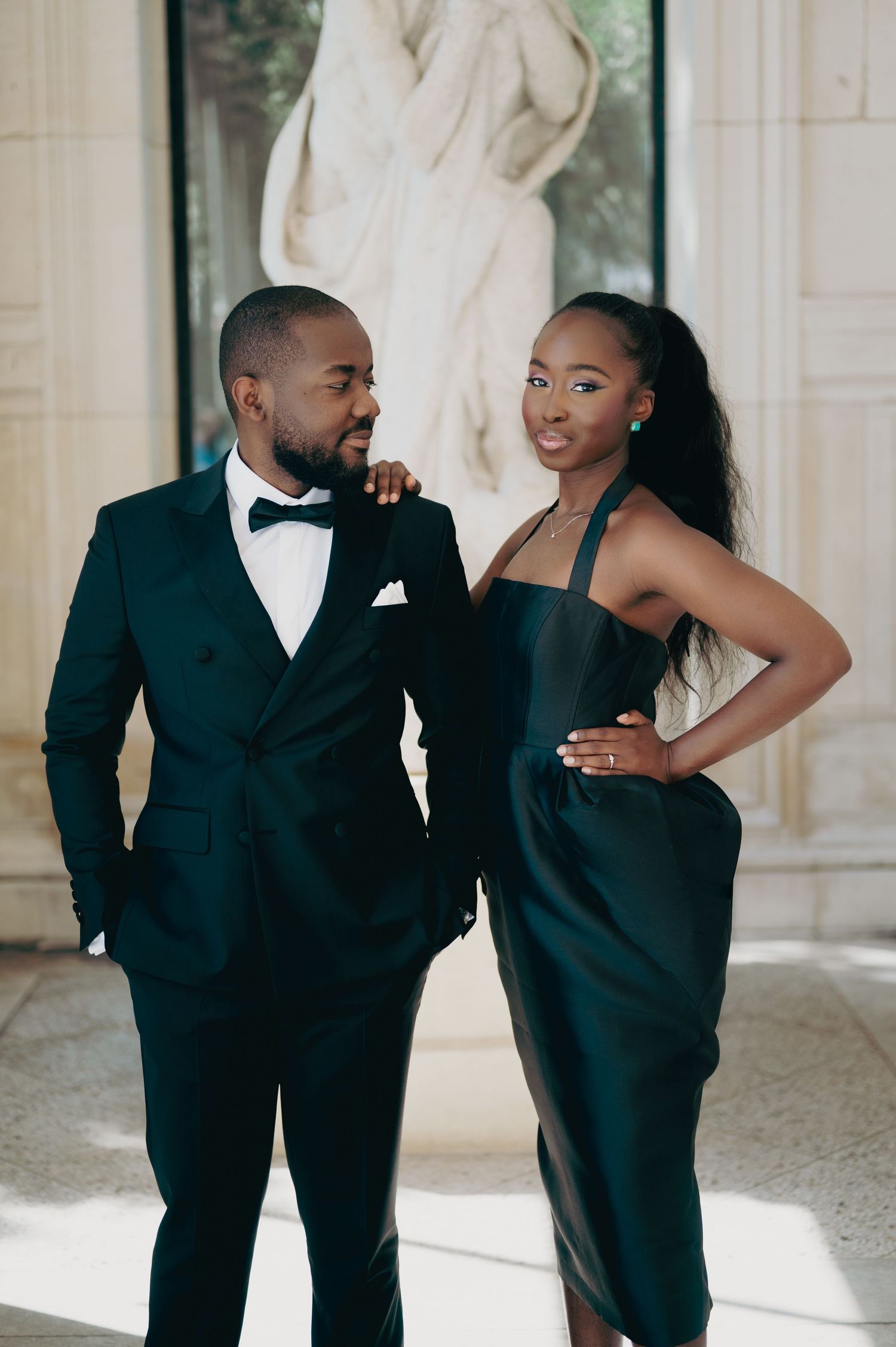 Fashionable couple posing in coordinated dark green formal outfits inside a classical architectural space with marble statue.