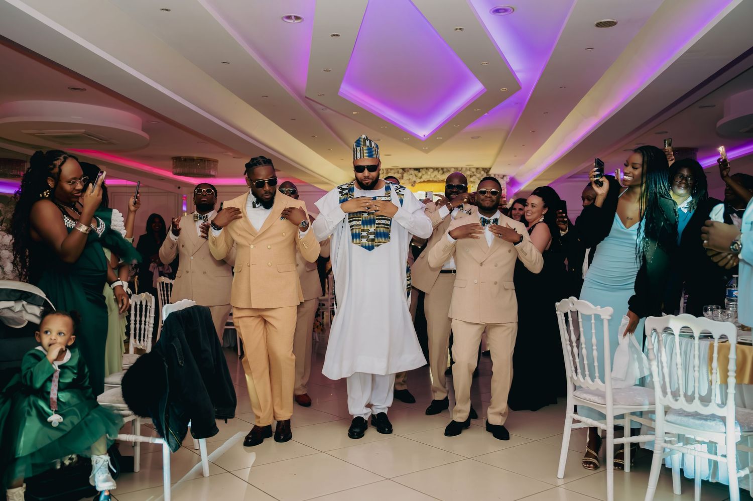 Groom in traditional African attire celebrates with groomsmen in cream suits at a wedding reception.