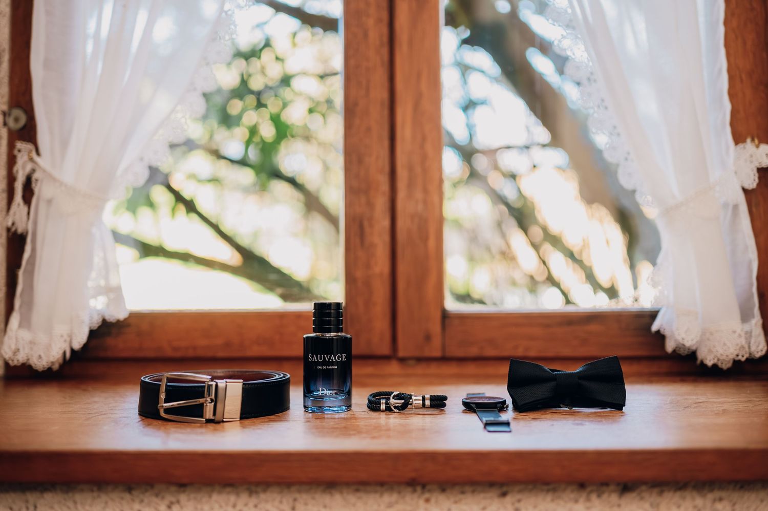 Groom accessories including cologne, belt, bow tie, and cufflinks arranged on a wooden windowsill.
