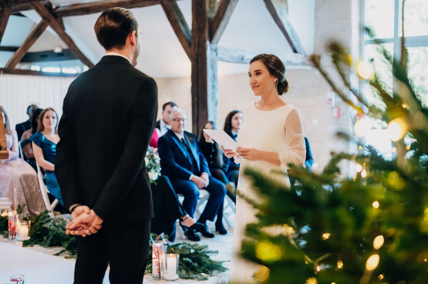 Smiling bride recites vows to groom as wedding guests watch during rustic barn ceremony.