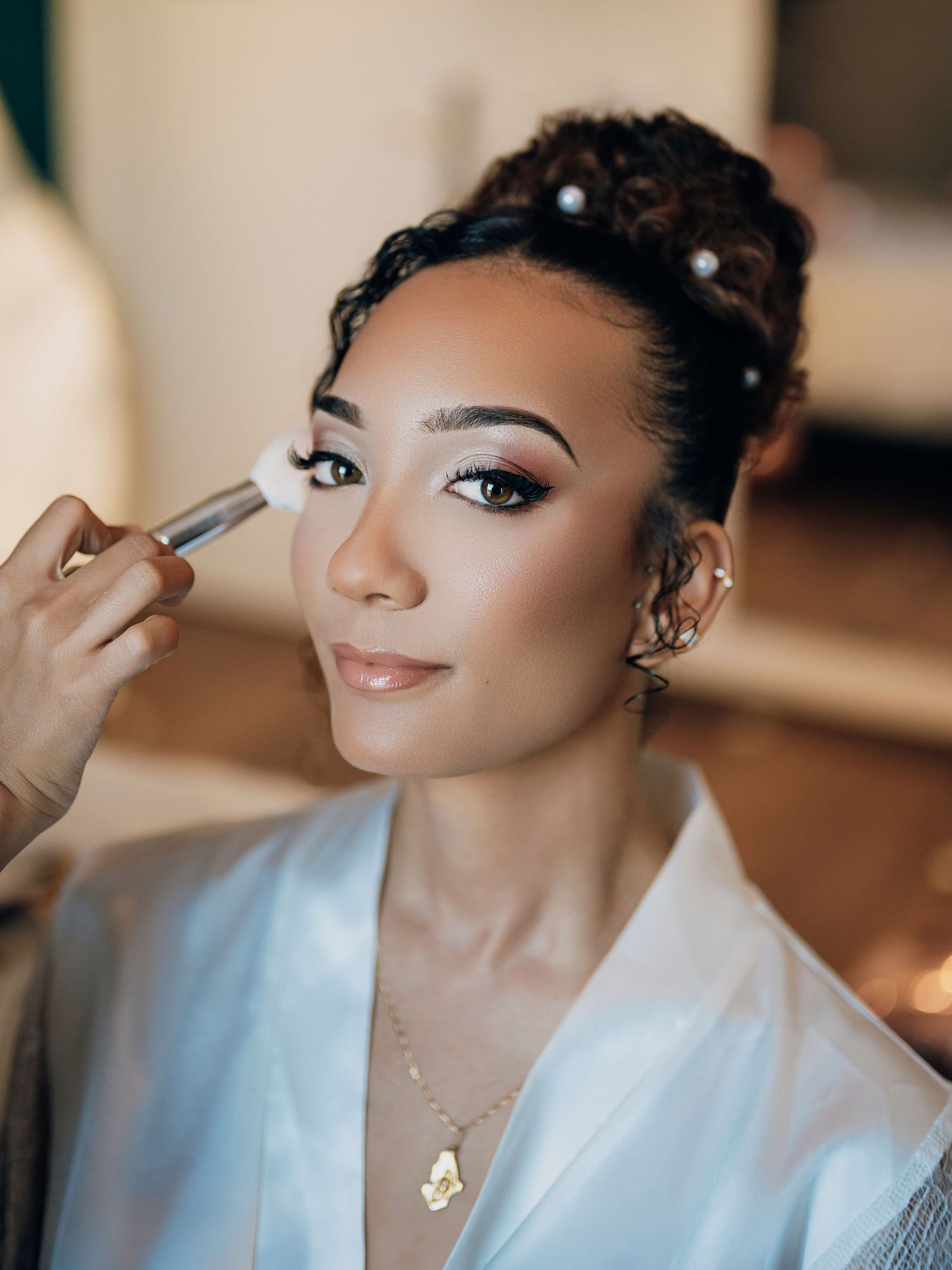 Beautiful bride getting makeup applied with brush while wearing white satin robe before wedding.