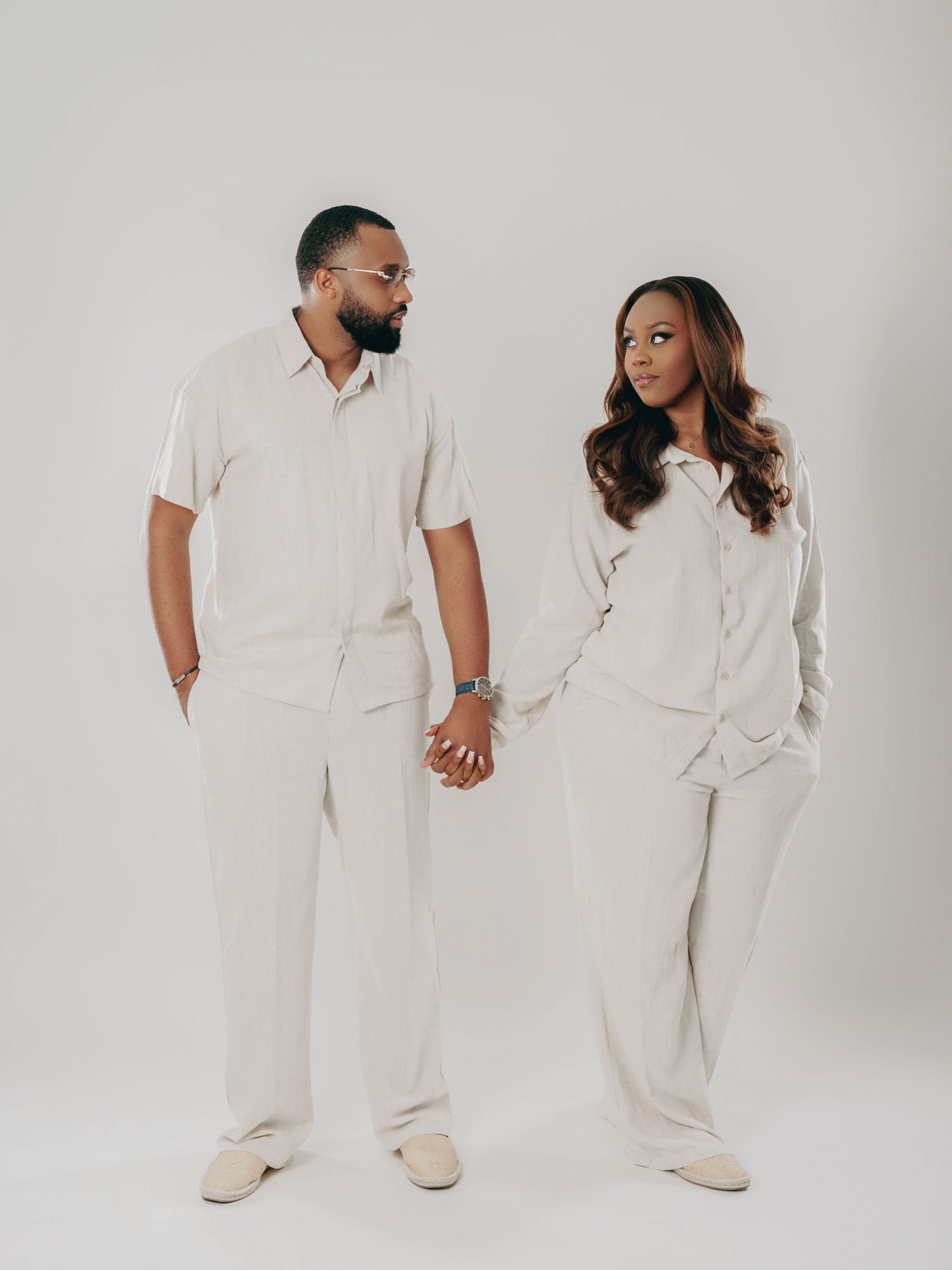 Stylish couple in coordinating cream linen sets hold hands while looking at each other in studio.