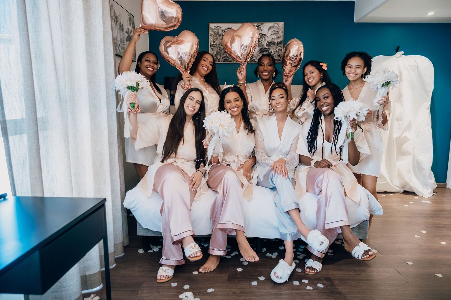 Bride posing with bridesmaids in matching robes holding rose gold heart balloons.