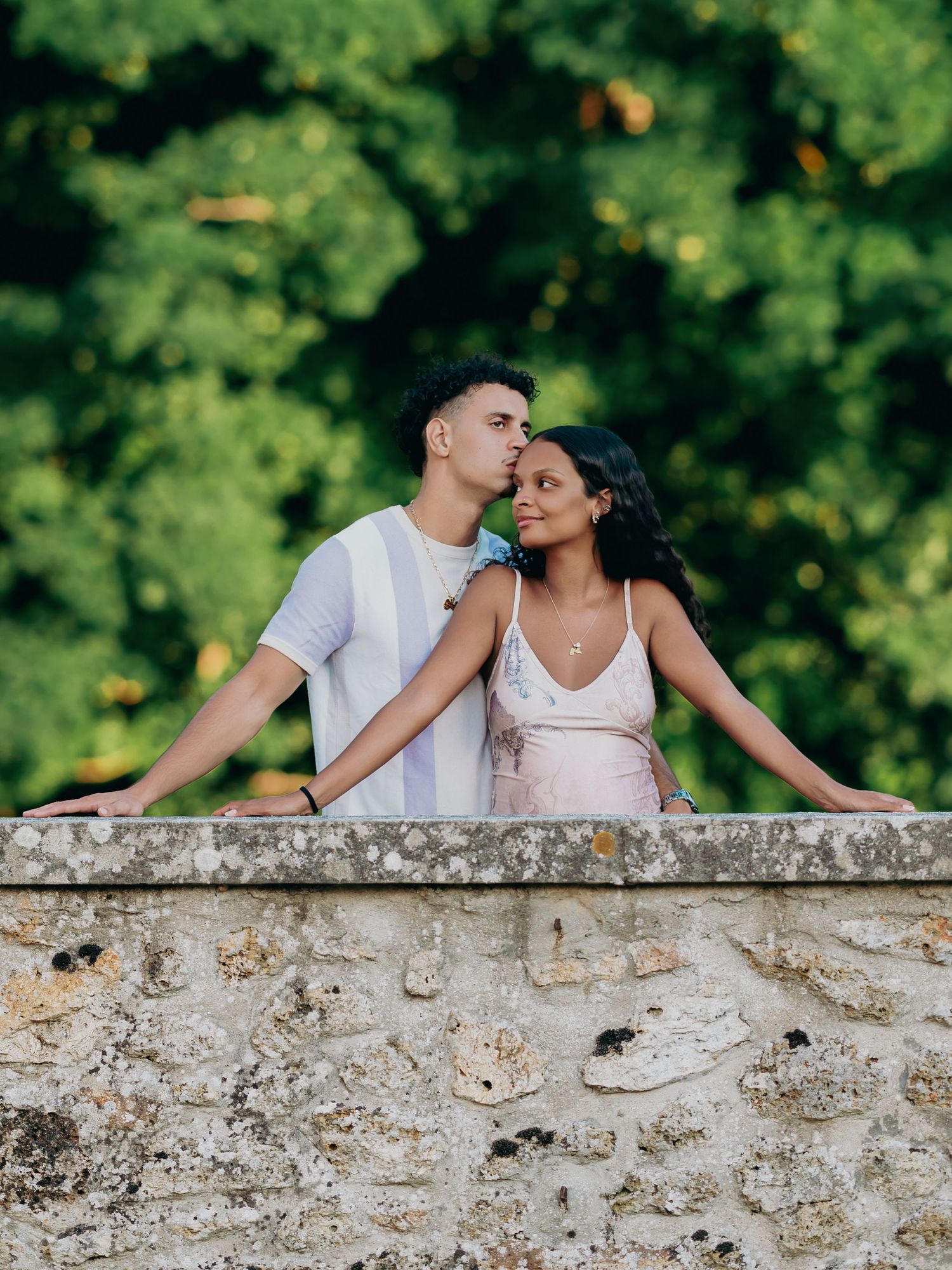 Couple sharing a tender kiss on a rustic stone wall surrounded by vibrant green trees.