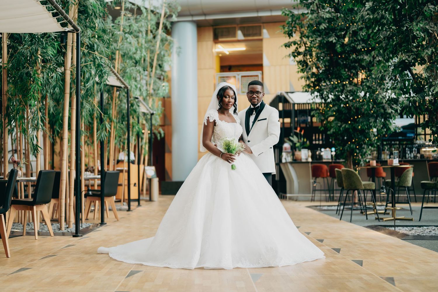 Elegant bride and groom posing in a modern venue surrounded by lush greenery on their wedding day.